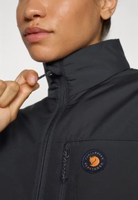 Svart jakke med glidelås og høy krage, med en oransje og marineblå sirkel logo på venstre side. Materialet ser glatt og lett ut.