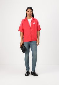 Camisa roja de manga corta con botones y logo, combinada con unos jeans ajustados azules y zapatos negros tipo slip-on. El modelo sostiene un bolso negro.