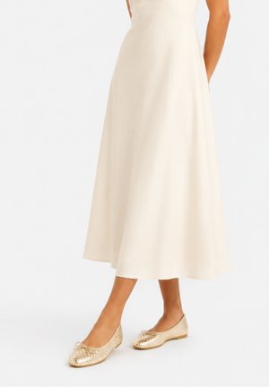 Donna con gonna midi color crema e ballerine dorate con texture, in piedi davanti a uno sfondo bianco semplice.