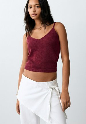 Donna che indossa un top senza maniche in maglia color bordeaux con spalline sottili e una gonna bianca a portafoglio legata in vita, in piedi davanti a uno sfondo semplice.