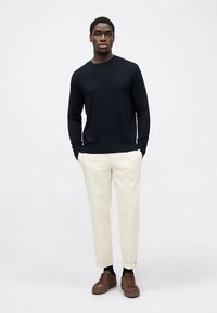 Tamsiai mėlynas megztinis, kreminės kelnės ir rudi sportiniai bateliai. Apranga pasižymi paprastu dizainu su apvalia apykakle ir pritaikyta forma.