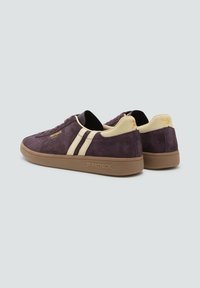 Zapatillas de ante moradas con rayas crema, suela de goma marrón y branding en el costado y el talón. Diseño de perfil bajo con un acabado texturizado.
