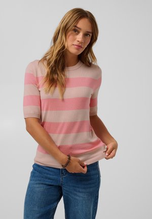 Ung kvinde med langt hår iført en kortærmet lyserød og beige stribet sweater og blå jeans, som holder i kanten af sin sweater.