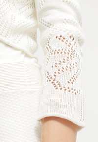 Vit långärmad stickad tröja med en texturerad kropp och öppen spetsdesign på ärmarna, som visar en delikat design och intrikata sömmar.