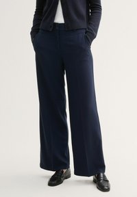 Marinefarbene Weitbeinerhosen aus einem glatten Stoff, mit einem Knopfverschluss, seitlichen Taschen und einer taillierten Silhouette. Schwarze Slipper runden den Look ab.