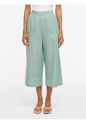 Žena v svetlozelených širokých krátkych nohaviciach s vysokým pásom, bielom topu a béžových žabkách, stojaca pred bielym pozadím.