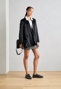 Blazer nero oversize, camicia bianca con colletto, gonna mini con fantasia e sneakers nere. Gli accessori includono una borsa nera con un accento dorato.