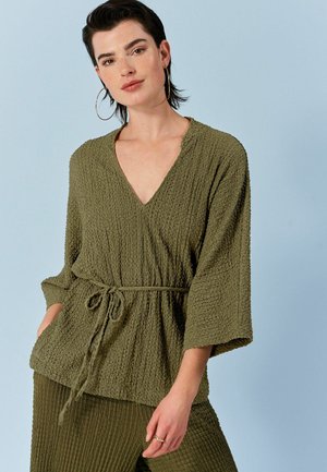 Olive rohelise, tekstuuriga ülemine osa V-kaelusega, kolmeveerand-svarrudega ja stringi taljega. Materjal näib olevat kerge ja kortsus.