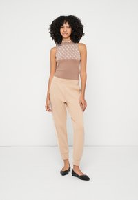 Beiges ärmelloses Oberteil mit gemustertem Netzstoff und hellbeige Jogginghose mit seitlichen Akzenten, kombiniert mit schwarzen Ballerinas. Glatte Texturen und tailliertes Design.