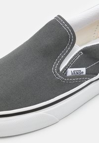 Chaussure grise à enfiler en toile avec des coutures blanches et une bande noire le long de la semelle. Présente une étiquette de logo Vans sur le côté.