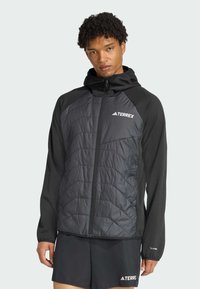 Un jeune homme aux cheveux bouclés porte une veste Terrex noire par-dessus une tenue sombre, se tenant devant un fond clair, regardant directement le spectateur.