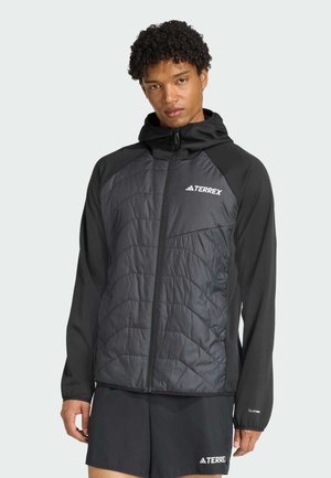 Een jonge man met krullend haar draagt een zwarte Terrex-jack over een donkere outfit, terwijl hij tegen een lichte achtergrond staat en recht in de camera kijkt.