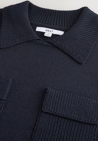 Strikket navy trøje med ribbet krave, der har to store teksturerede klaftlommer og et mærke med teksten "NEXT."