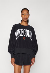 Zwarte fleece sweatshirt met "NIKECOURT" in witte vetgedrukte letters, ribbelboord bij de hals, en bijpassende shorts met een kleurrijke logo patch.