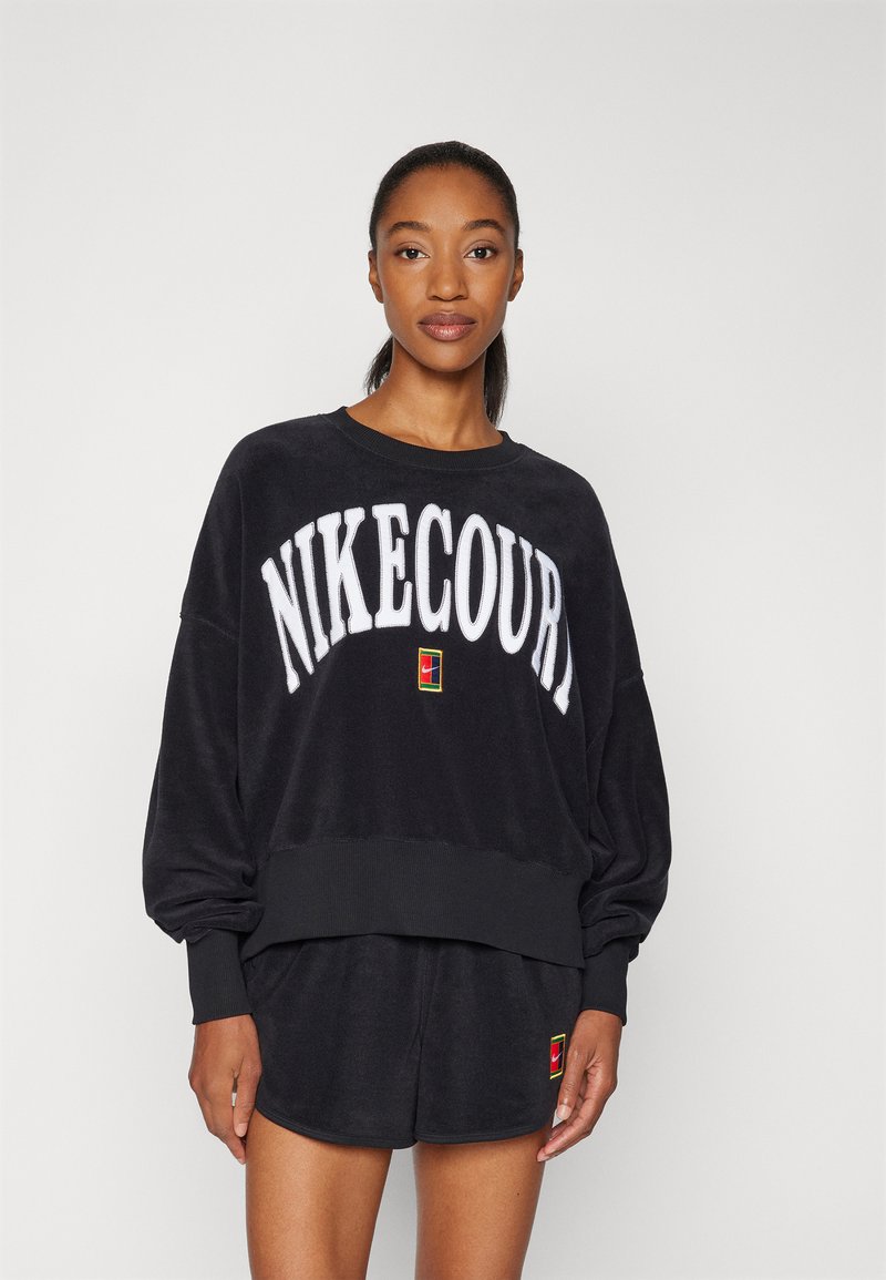 Zwarte fleece sweatshirt met "NIKECOURT" in witte vetgedrukte letters, ribbelboord bij de hals, en bijpassende shorts met een kleurrijke logo patch.