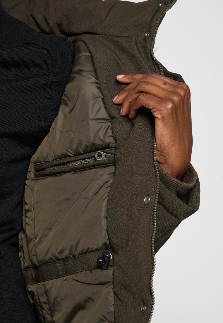 Doudoune olive avec doublure matelassée, présentant une poche zippée et une coque extérieure texturée. Boutons sur le devant.