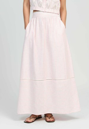 Femme portant une jupe longue taille haute rayée rose et blanche avec des poches, ainsi que des sandales marron à lanières, sur un fond uni.