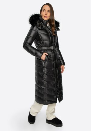 Vrouw draagt een lange zwarte gewatteerde jas met riem en bontkraag, witte broek en zwarte instapschoenen, staand met handen in de zakken.