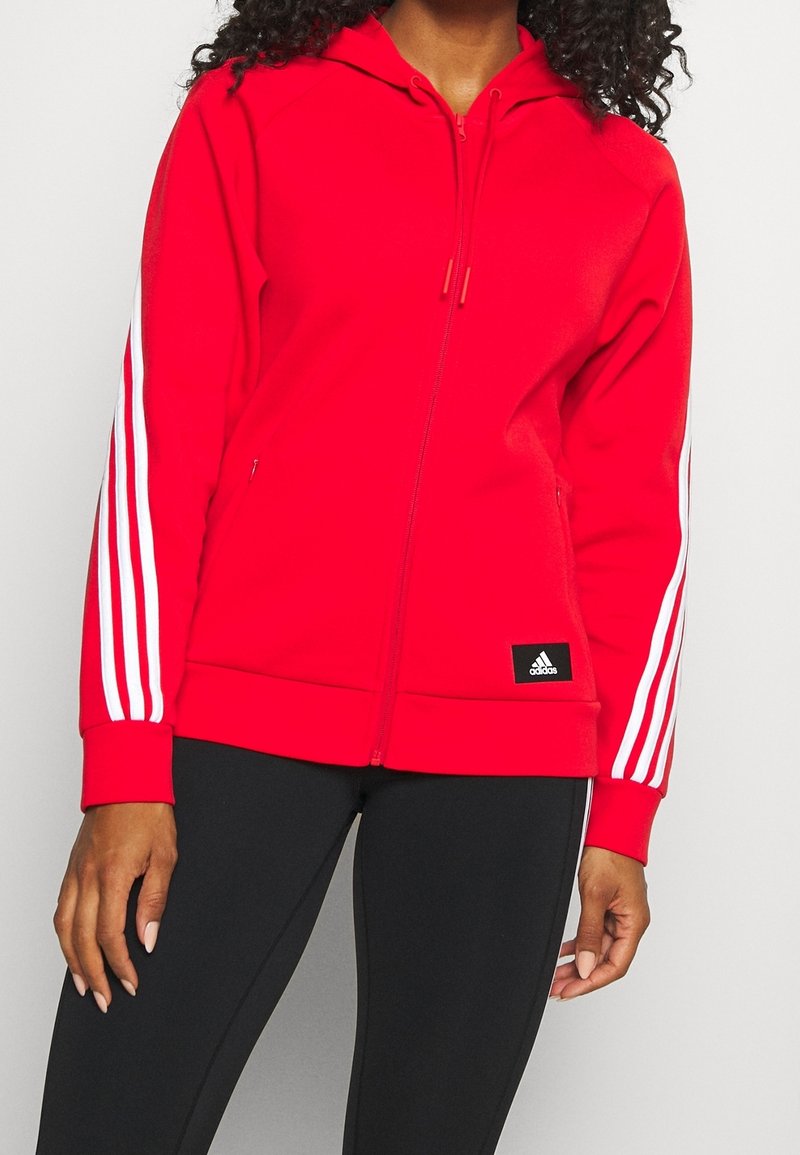 Sudadera roja con cremallera, fabricada en tela suave y con capucha ajustable, que presenta acentos en blanco con tres rayas en las mangas y un logo de Adidas en la parte inferior izquierda.
