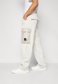 Béžové cargo kalhoty s boční kapsou, vyrobené z pevné látky. Mají přímý střih a elastické manžety. Doplněny bílými teniskami.