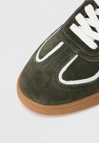 Grüne Wildleder-Sneaker von vorne mit weißen Schnürsenkeln, weißen Steppnähten und einer hellbraunen Gummisohle auf weißem Hintergrund.