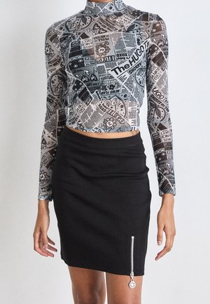 Kobieta ubrana w długi crop top z nadrukiem gazetowym i czarną mini spódniczkę z pionowym zamkiem błyskawicznym oraz zawieszką uśmiechniętej buźki.