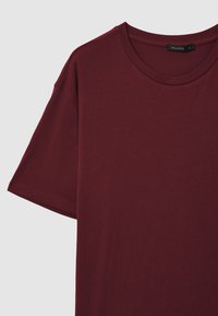 T-shirt in cotone bordeaux a maniche corte con scollo rotondo, cuciture visibili sulle maniche e una piccola etichetta nera con la taglia all'interno del colletto.
