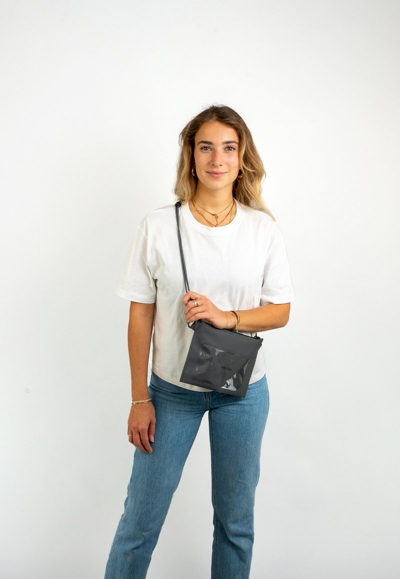 Bolso bandolera negro con bolsillo frontal transparente, forma rectangular, textura suave y correa ajustable, sostenido por una persona con atuendo casual.