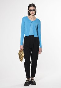 Hellblauer Cardigan über einem passenden Oberteil, kombiniert mit schwarzen Cargo-Hosen. Die Accessoires umfassen eine goldene Clutch und schwarze klobige Schuhe.