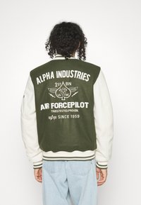 Giacca varsity con lana verde oliva e maniche sintetiche color crema. Presenta grandi scritte bianche e grafiche ispirate al mondo militare sul retro.