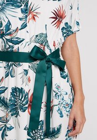 Robe blanche à imprimé feuilles et fleurs tropicales vertes, bleues et rouges, cintrée à la taille avec une ceinture en satin vert foncé nouée en nœud.