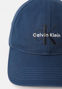 Tamsiai mėlyna beisbolo kepuraitė, pagaminta iš tekstūrinio audinio, su siuvinėjamu "Calvin Klein" logotipu ir "CK" kontrastingomis juoda bei balta spalvomis.