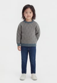 Bambino con capelli a media lunghezza che indossa un maglione a quadri, jeans blu e scarpe da ginnastica bianche, in piedi davanti a uno sfondo semplice.