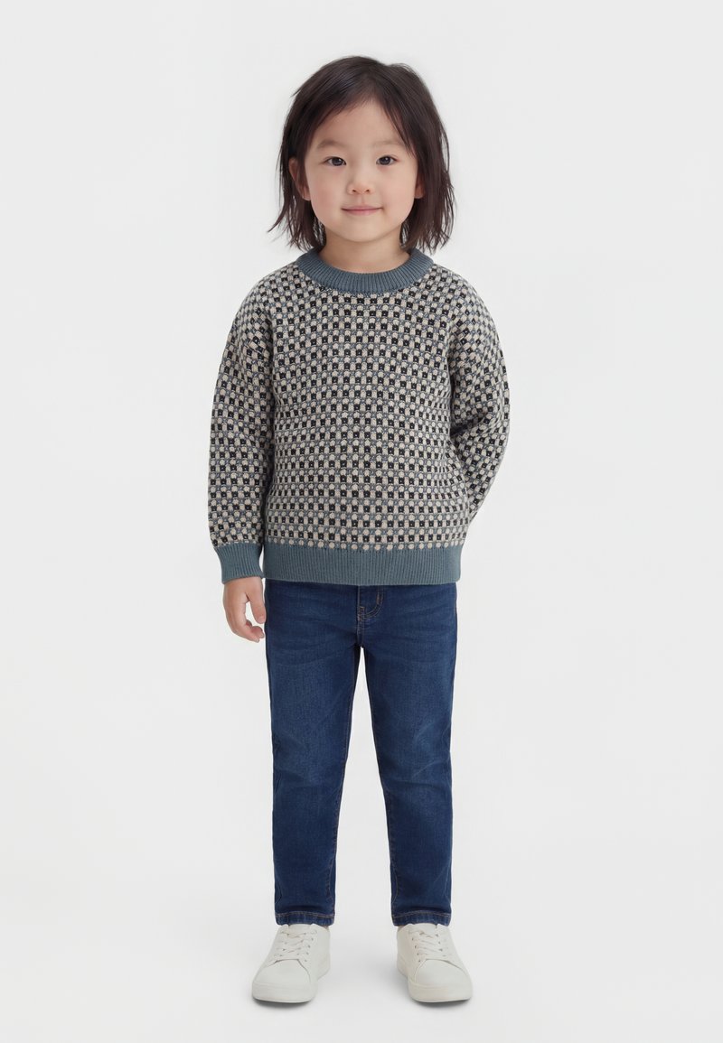 Bambino con capelli a media lunghezza che indossa un maglione a quadri, jeans blu e scarpe da ginnastica bianche, in piedi davanti a uno sfondo semplice.