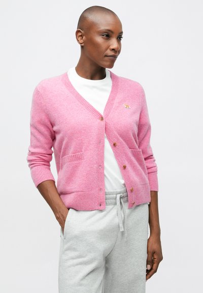 Cardigan roz, fabricat dintr-un material moale, cu decolteu în V, închidere cu nasturi și două buzunare frontale, purtat cu pantaloni sport gri.