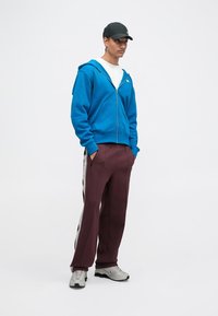 Sudadera azul con cremallera, camiseta blanca debajo, pantalones de chándal burdeos con franjas laterales, zapatillas deportivas grises y una gorra negra.