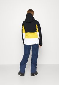 Veste de ski en blocs de couleur avec des panneaux noirs, jaunes et blancs, dotée d'une capuche et d'une texture en tissu doux ; associée à un pantalon bleu et des bottes noires.