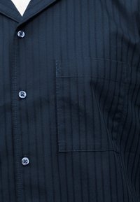 Μαύρο μπλε πουκάμισο με γιακά και κάθετες ρίγες, τρία ορατά κουμπιά και τσέπη στο στήθος στην αριστερή πλευρά.