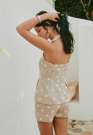 Sötét hajú nő bézs mintás nyakpántos felsőben és rövidnadrágban haját igazítja fehér fal és kőpadló előtt.