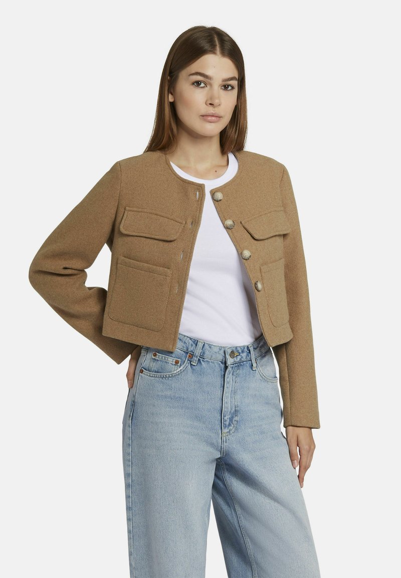 Geknipte, lichtbruine wollen jas met knoopsluiting en twee voorkezen; gedragen over een witte top en gecombineerd met lichtblauwe jeans.