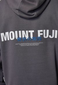 Sweat à capuche gris arborant "MONT FUJI" en grandes lettres blanches et du texte japonais, avec des détails en broderie bleu et un tissu texturé.
