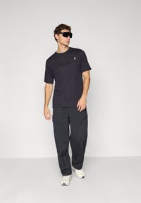 Black short-sleeve T-shirt with a subtle pattern on the chest, paired with loose-fitting black cargo pants and light grey sneakers.