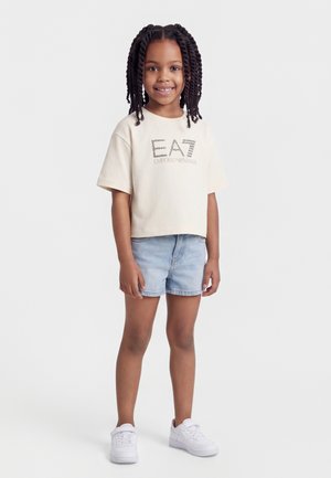 Fille souriante aux cheveux tressés portant un t-shirt beige EA7, un short en jean clair et des baskets blanches, se tenant devant un fond uni clair.