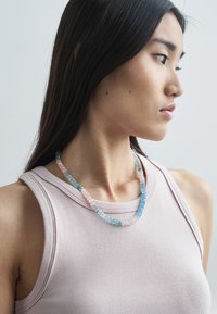 Perlekæde med pink, blå og gennemsigtige sten i forskellige former, trukket på en snor, båret sammen med en blød, lysefarvet tanktop.