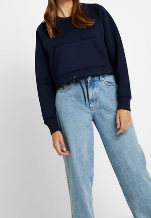 Femme portant un sweat-shirt court bleu marine avec poche avant et un jean taille haute bleu clair, debout devant un fond uni.