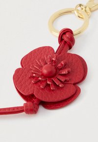Sarkans ādas atslēgu piekariņš, kas veidots kā zieds ar mezglotu stublāju, pievienots zelta metāla atslēgu gredzenam uz baltas fona.