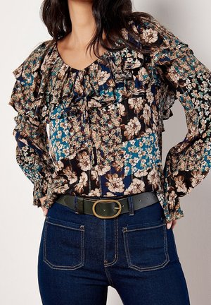 Kvinde iført en bluse med blomsterprint og flæser, stoppet ned i mørkeblå jeans med et sort bælte med en messing oval spænde.