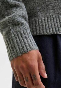 Grå strikket sweater med ribbede manchetter, viser et nærbillede af en hånd med synlige tatoveringer, sat mod en mørk stofbaggrund.