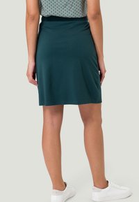 Grüner Skater-Rock aus weichem Stoff, mit einer taillierten Taille und glatter Textur. Kombiniert mit weißen Sneakers und einem gemusterten Oberteil.