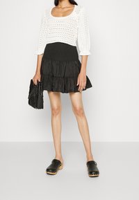 Vrouw in een witte gehaakt top met driekwart mouwen, een zwarte gelaagde minirok, zwarte klompen met houten zolen en een zwarte franjerugtas.